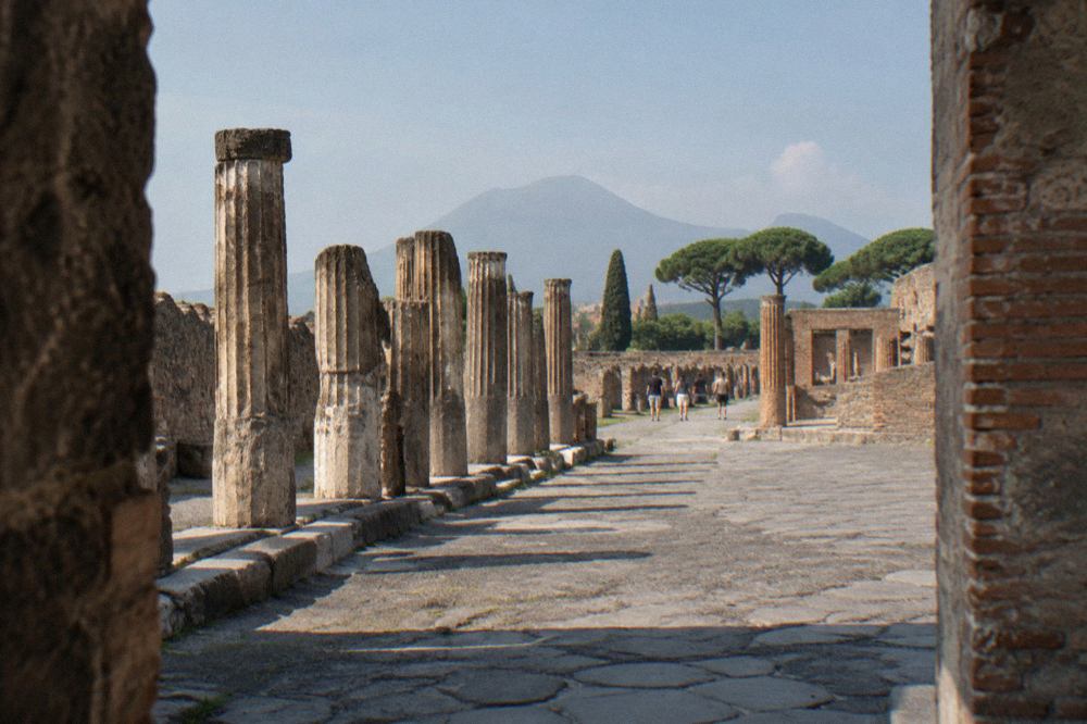 The excavated stone street of ancient Pompeii with Mount Vesuvius rising behind it under a pale sky