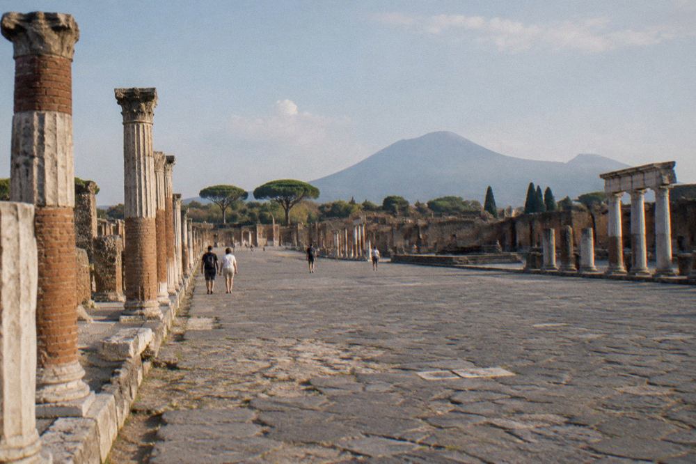The excavated stone streets of Pompeii with the green slopes of Mount Vesuvius rising behind the ruins under a clear sky