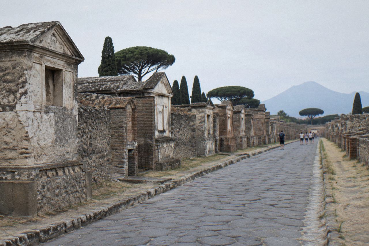Sunlit stone street in the excavated Roman city of Pompeii with ancient walls and Mount Vesuvius on the horizon.