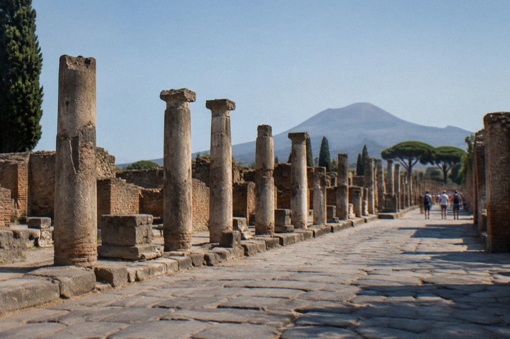 Excavated stone walls and columns of Pompeii with grassy unexcavated ground rising beyond them