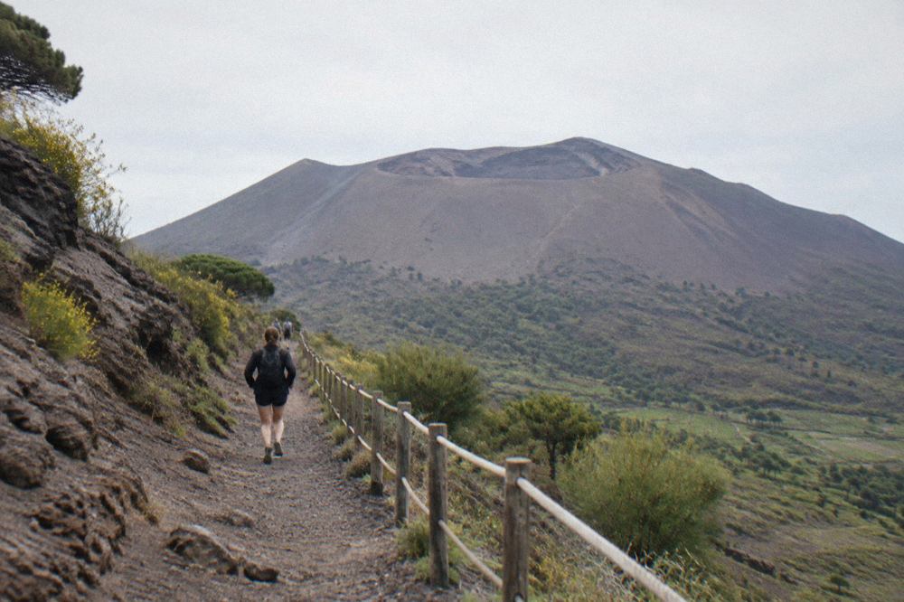 The rocky rim of the Vesuvius crater under a wide grey sky, with dark volcanic slopes falling away to the Bay of Naples