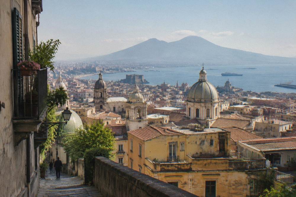 A sunlit Naples street with old buildings and a distant view over the Bay of Naples toward a volcano