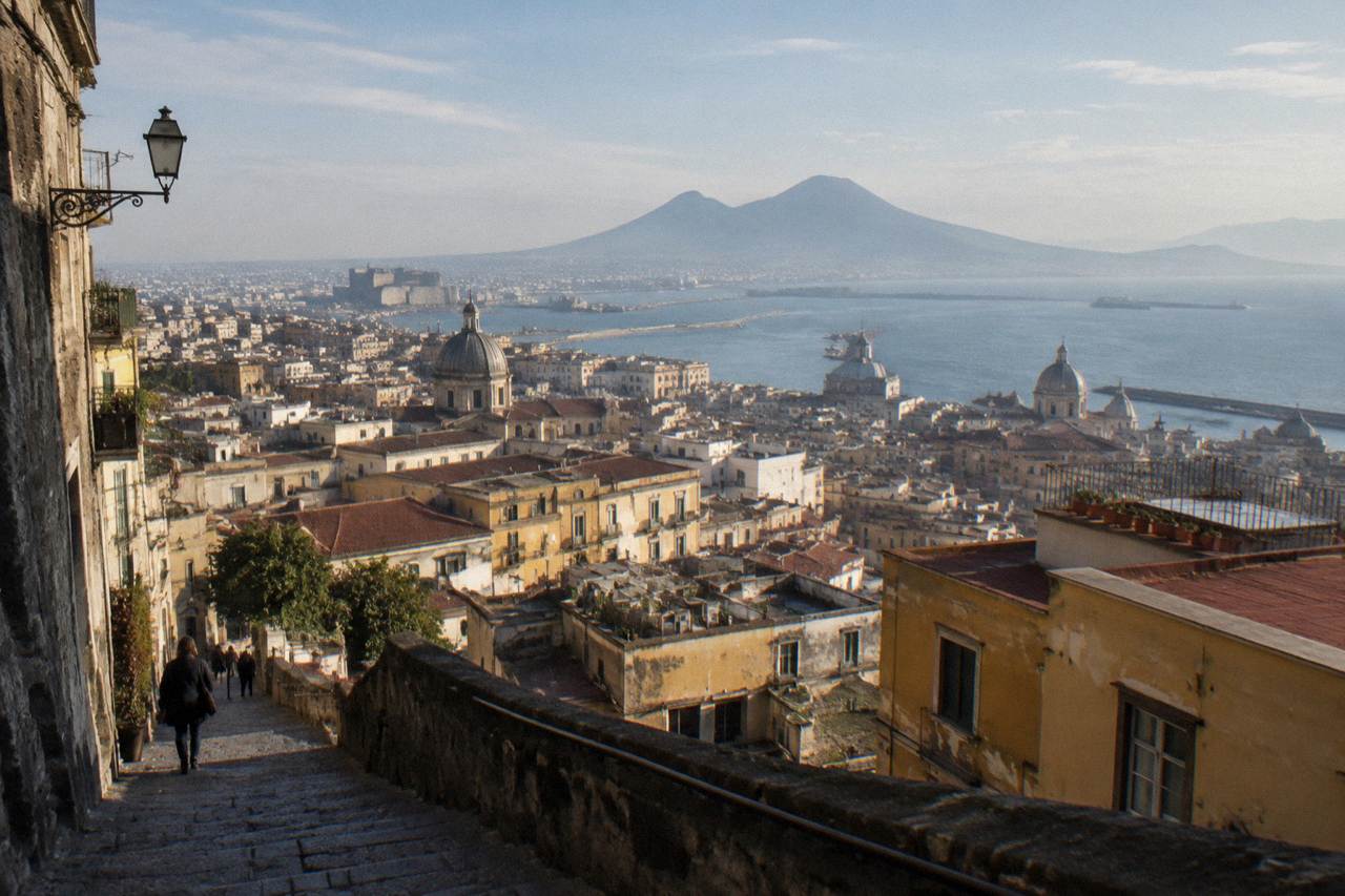A sunlit Naples street with old buildings and a distant view over the Bay of Naples toward a volcano