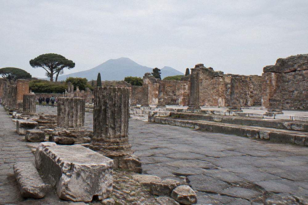 Excavated Roman stone walls of Pompeii with grassy unexcavated mounds rising behind them under a clear sky