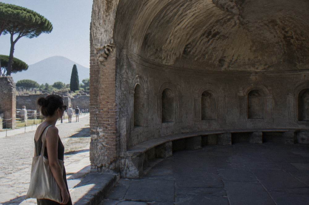 A Roman villa terrace with stone columns looking out over the blue Bay of Naples toward a distant volcano