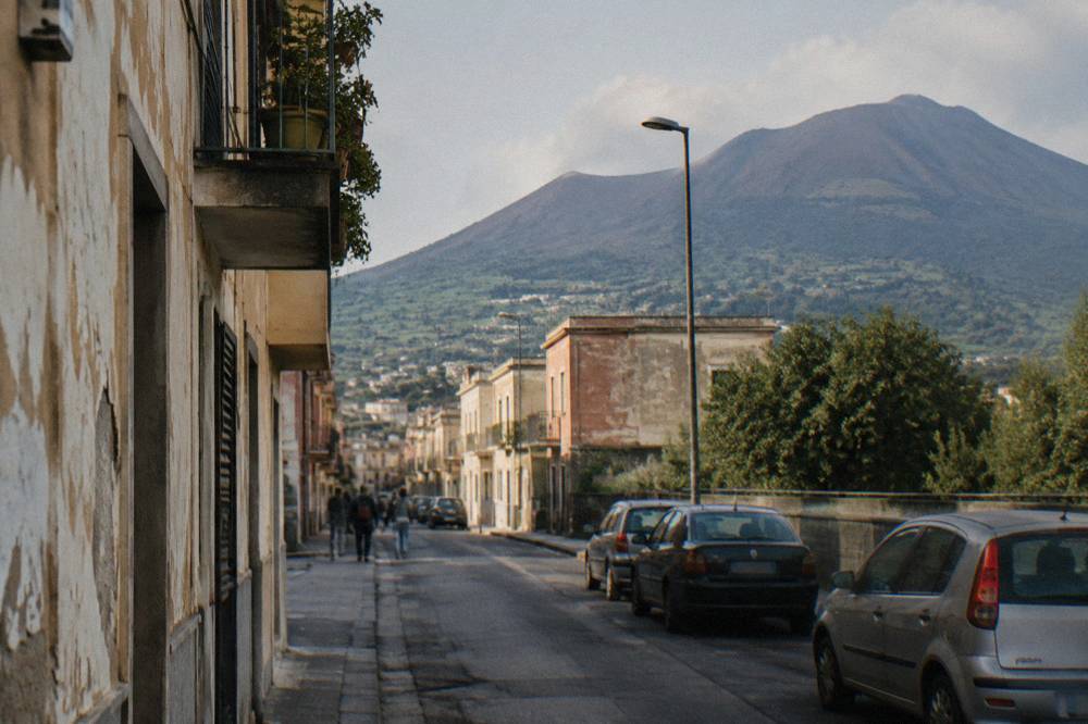 An ancient Roman street of stone paving running between ruined walls toward a distant volcano cone