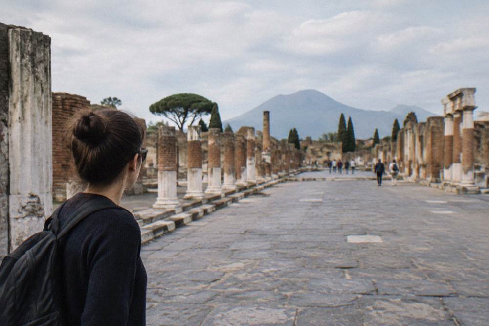The open rectangular forum square of ancient Pompeii with stone paving and column bases under a bright sky, Mount Vesuvius on the horizon
