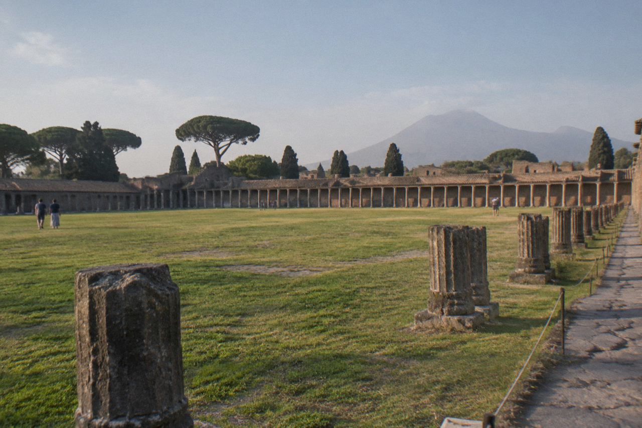 A grassy rectangular field framed by low portico walls under plane trees inside an ancient Roman archaeological site