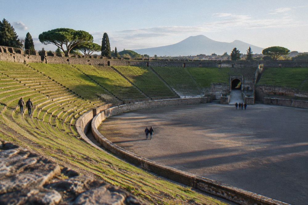 The grassy interior arena of the ancient Pompeii amphitheatre ringed by stone seating tiers under a clear sky