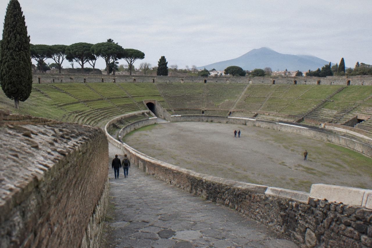 The grassy interior arena of the ancient Pompeii amphitheatre ringed by stone seating tiers under a clear sky