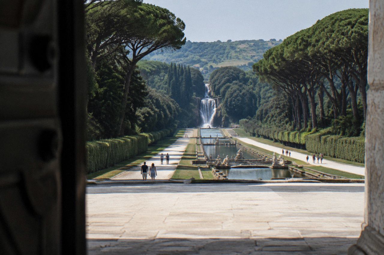 A grand Baroque palace facade with a wide forecourt and tree-lined avenue under a clear sky