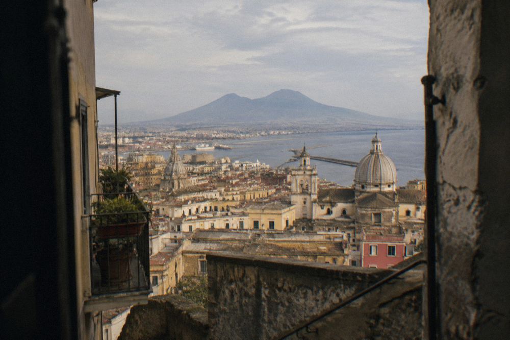 A busy street in the historic centre of Naples with tall buildings, balconies and a pizzeria on the corner