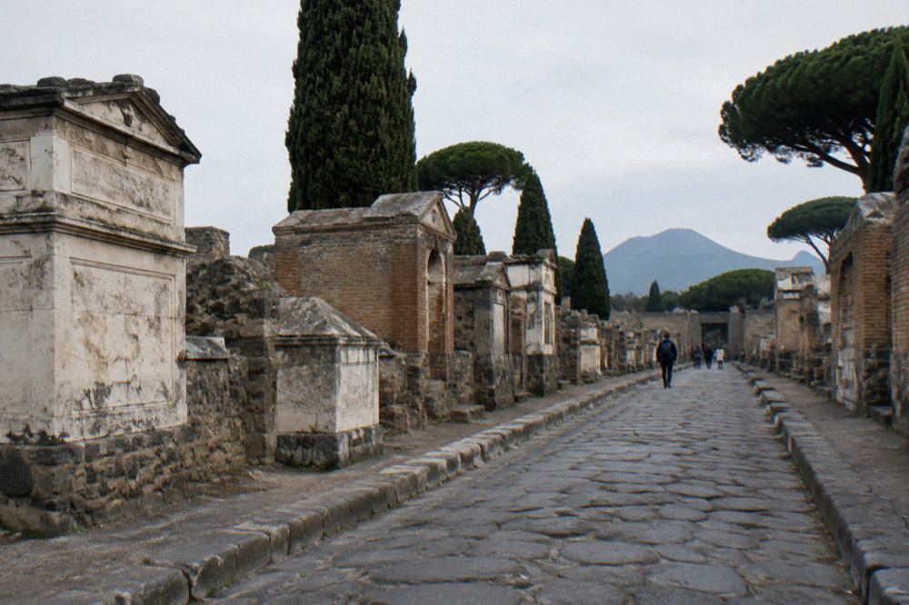 Wide empty street in Pompeii lined with stone walls and Mount Vesuvius in the distance