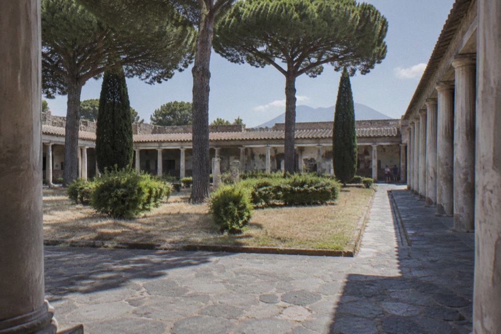 A quiet paved street with stone walls and an entrance arch to an archaeological site in bright morning sun