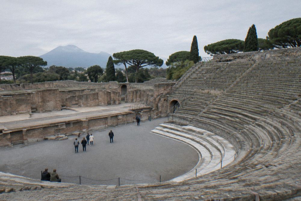 A small group of visitors listening to a guide in the Forum of Pompeii with the Temple of Jupiter behind them