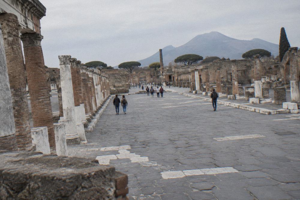 Crowded stone pathway in Pompeii with visitors walking between ancient ruined buildings in warm light